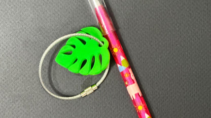Cute personalized small monstera leaf in bright green with a 2mm silver coated cable wire keychain next to a mechanical pencil for size comparison.